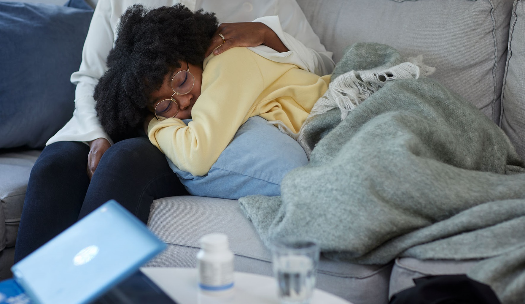 En flicka ligger och vilar med slutna ögon under en filt. Flickan har huvudet i sin förälders knä, föräldern håller om flickan med ena handen. I förgrunden finns ett bord med en laptop, diverse förpackningar med värktabletter samt ett glas vatten.