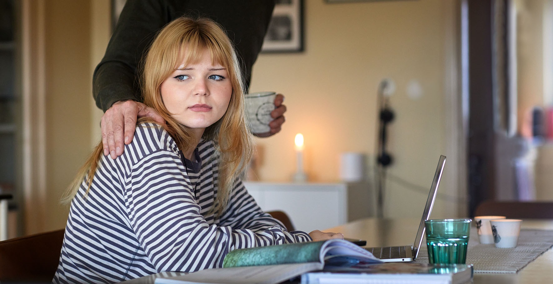 Barn som sitter i hemmamiljö och studerar men ser ut att ha svårt att fokusera. Förälder står snett bakom med en hand på axel och böjer sig fram som för att kolla om allt är okej.